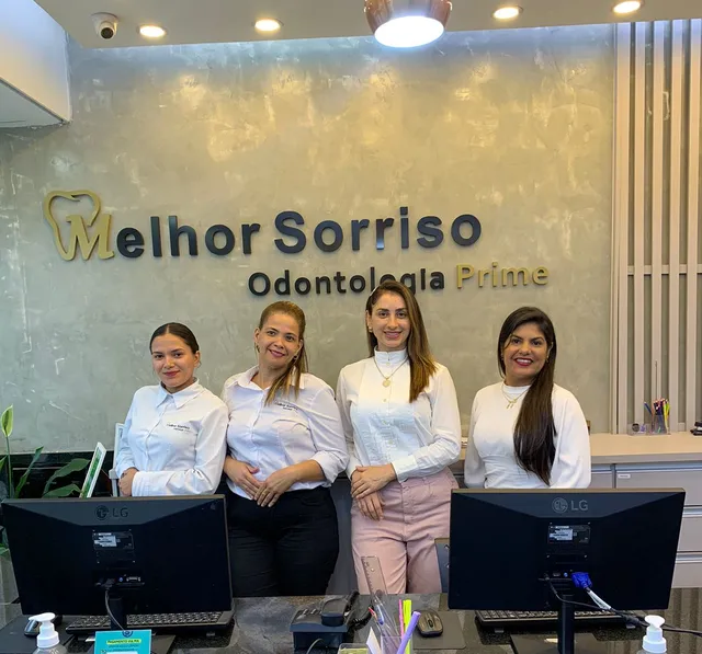Quatro integrantes da equipe de recepção, de camisa branca, atrás do balcão com o letreiro da clínica ao fundo